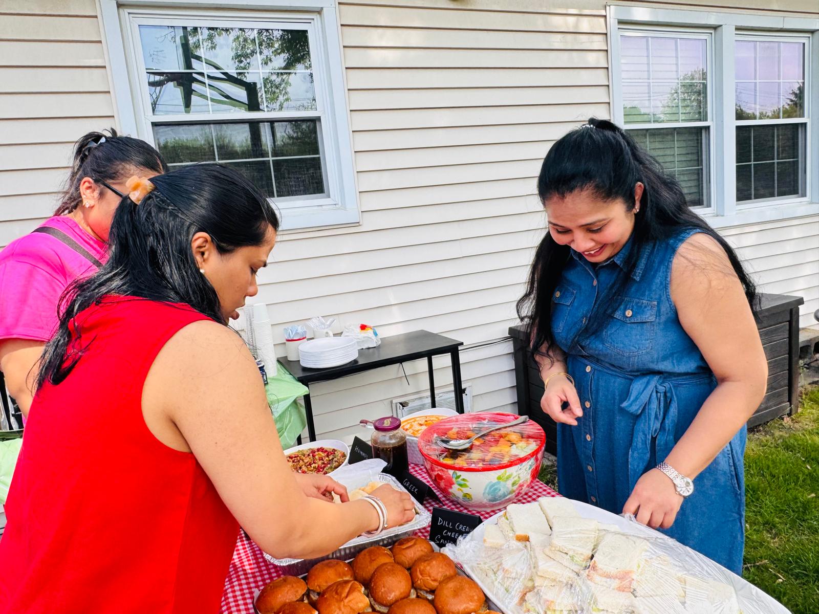 Three women setting up the food table with sliders, sandwiches, fruit salad, hummus and chaat on red gingham