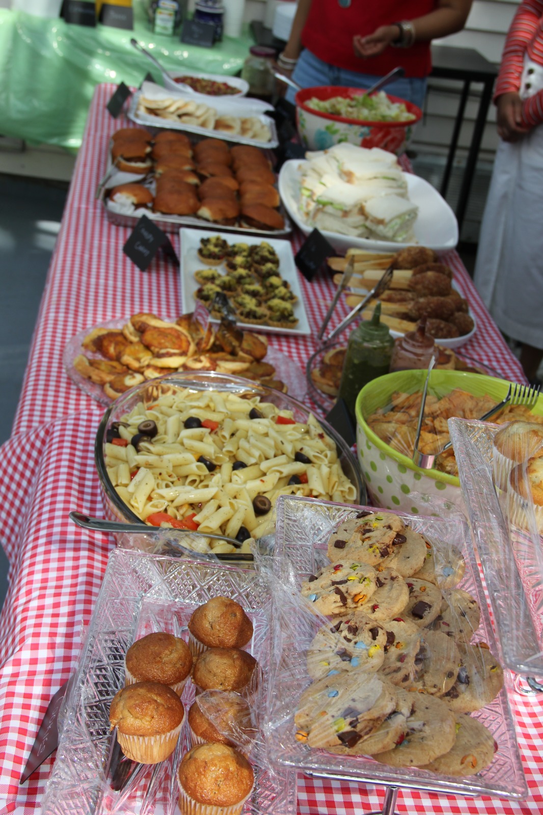 Long buffet table with sandwiches, pasta salad, cookies, muffins and other homemade dishes on red gingham tablecloth