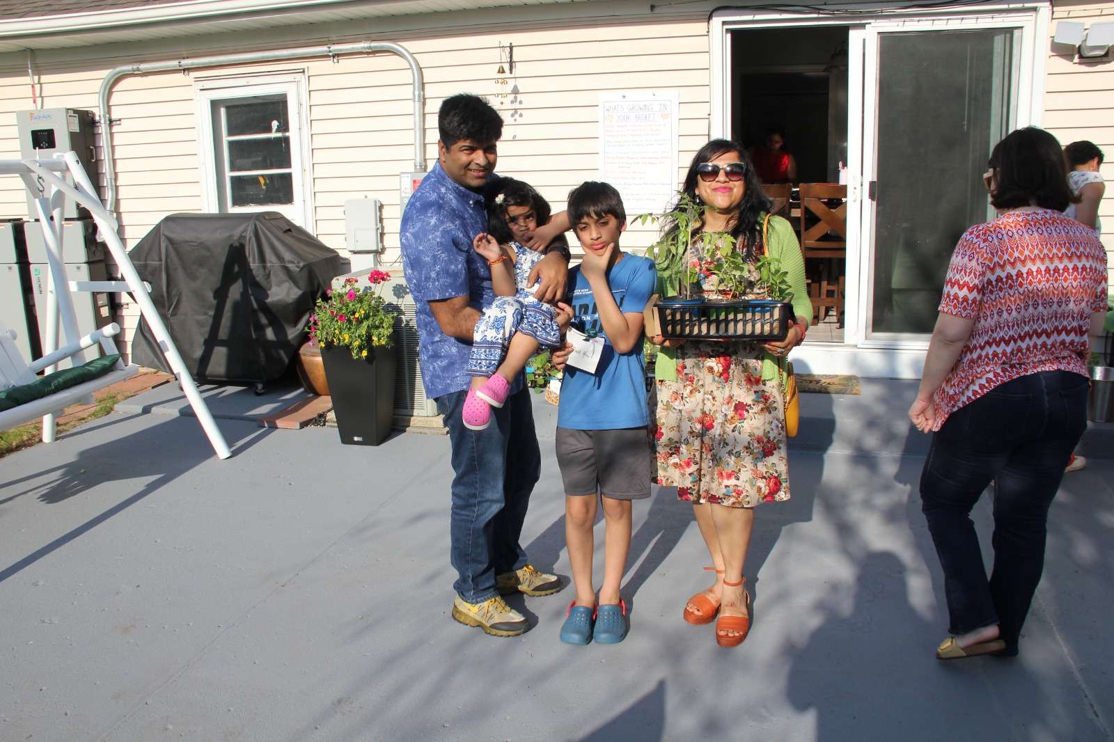 Family with young kids holding a tomato plant giveaway basket on the patio under the basket menu board
